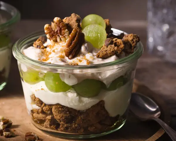 Ein Glas mit cremigem Dessert, geschichtet mit grünen Trauben, zerbröselten Keksen und Nüssen. Ein köstliches Trauben-Cookie-Dessert, perfekt für jeden Anlass.