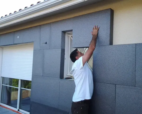 Un ouvrier pose des panneaux isolants gris pour l'isolation par l'extérieur d'une maison, près d'une fenêtre avec volet roulant.