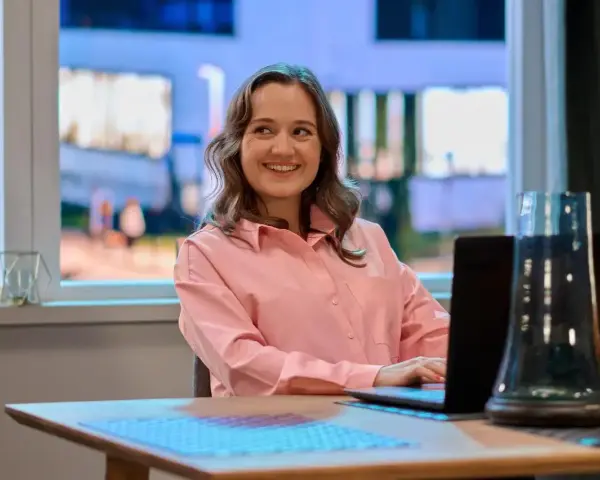Uśmiechnięta Dorota Krempa pracuje przy laptopie, siedząc przy stole. W tle widać okno z nocnym widokiem.