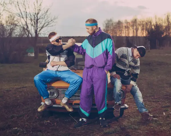 Trzech mężczyzn w strojach z lat 90. pozuje na tle starego samochodu. Jeden z nich ma na sobie fioletowy dres, drugi biało-czarną kurtkę i niebieskie spodnie, a trzeci sweter w geometryczne wzory.