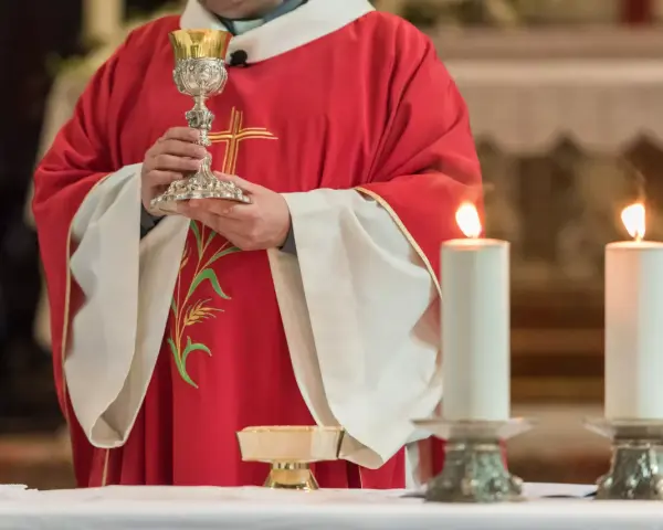Kapłan w czerwonej szacie z krzyżem trzyma kielich podczas konsekracji. Płoną dwie świece.