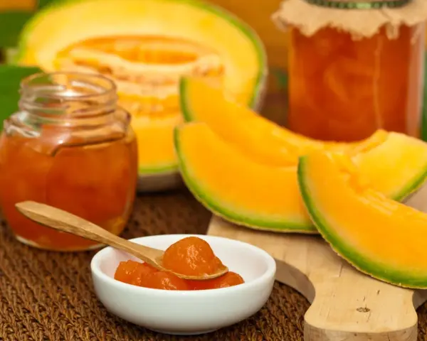 Marmellata di melone in vasetti, con fette di melone fresco e un cucchiaino pieno di confettura.