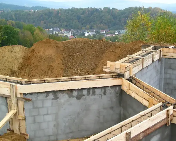 Fundamenty domu w budowie, zbrojenie i szalunki. Widać kopce ziemi i zabudowania w tle. Ważne jest pozwolenie na budowę, by wszystko szło zgodnie z planem.