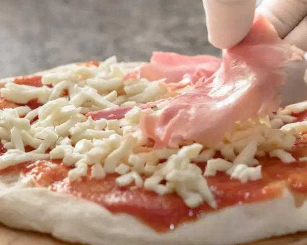 Ręka w białej rękawiczce kładzie plasterki szynki na pizzy z serem i sosem pomidorowym.