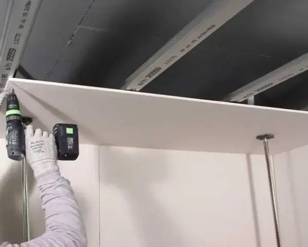 Persona con guanti avvita una lastra di cartongesso al soffitto metallico, un progetto di cartongesso fai da te.