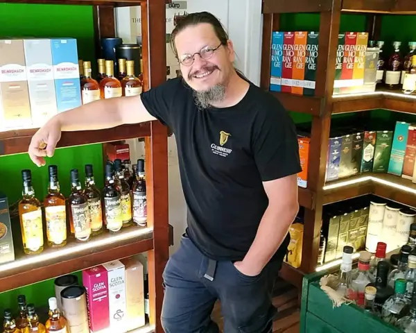 Mann in einem Guinness-T-Shirt steht in einem Laden voller Whisky-Flaschen und -Schachteln. Er lächelt und lehnt sich an ein Regal.