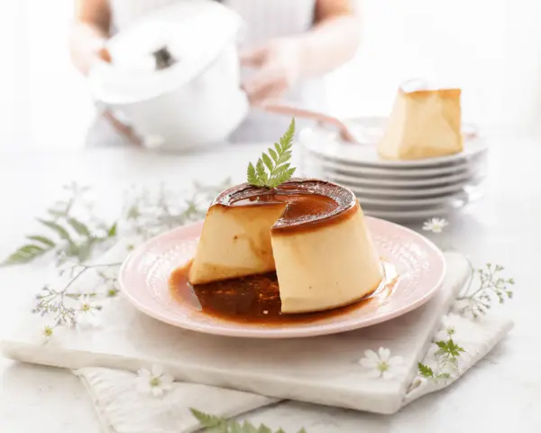 Delicioso flan de queso con caramelo líquido, decorado con una hoja verde. En el fondo, una persona prepara más postres.