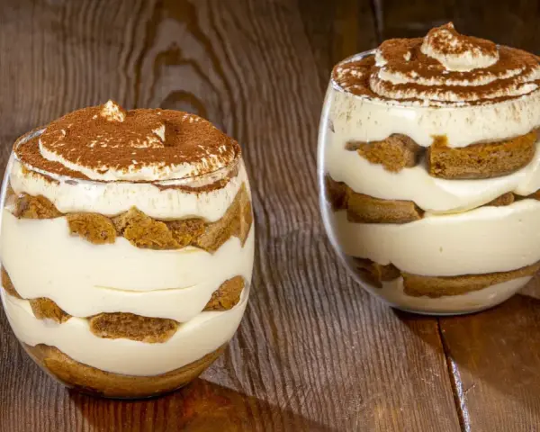 Due bicchieri di tiramisù senza uova, stratificati con crema e savoiardi, spolverati di cacao.