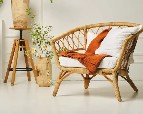 Un fauteuil en rotin tressé, invitant à la détente. Un plaid orange et un coussin blanc ajoutent une touche de confort. L'entretien rotin est essentiel pour préserver sa beauté naturelle.