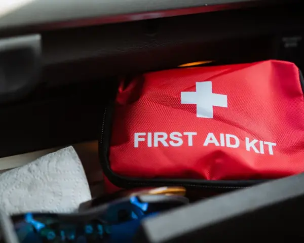 Czerwona apteczka samochodowa z białym krzyżem i napisem "FIRST AID KIT" leży w schowku. Obok niej papierowy ręcznik.