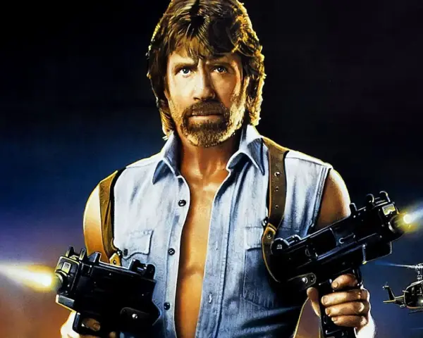 Chuck Norris z karabinami w akcji, jak w jego najlepszych filmach. W tle helikoptery i budynek.
