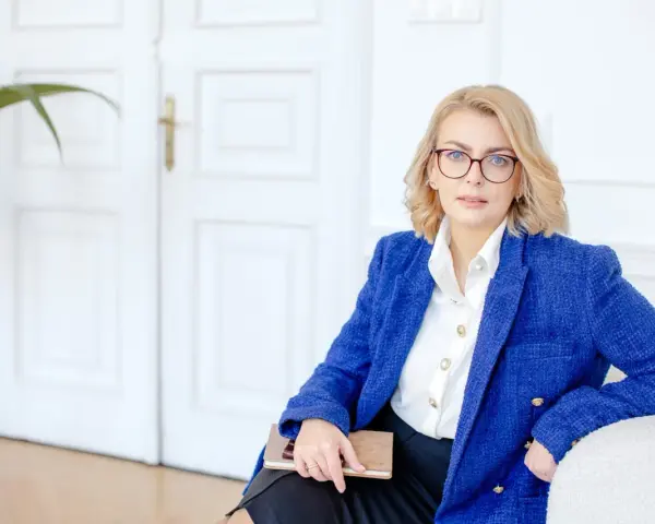Kobieta w okularach i niebieskiej marynarce trzyma notatnik. Planuje dofinansowanie na otwarcie firmy.