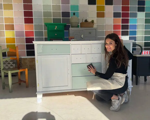 Mujer sonríe mientras lija un mueble para pintar. Detrás, un muro con muestras de colores de pintura.