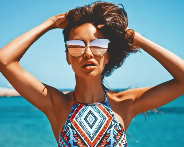Kobieta w okularach przeciwsłonecznych na plaży, z rękami w gorze. Jej skóra ma piękny, złocisty odcień, jakby świadczył o wspaniałych beta karoten efektach.