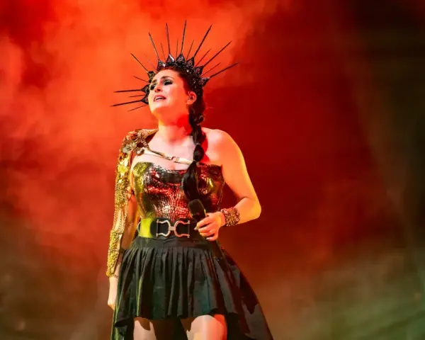 Wokalistka Within Temptation w koronie i błyszczącej kreacji, z mikrofonem w dłoni, na tle czerwonej mgły.