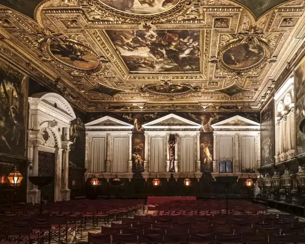 Sala capitolare Scuola Grande di San Rocco: soffitto dorato affrescato, pareti con dipinti e statue, sedie rosse disposte per un evento.
