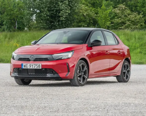 Czerwony Opel Corsa E na tle zieleni. Zbliżenie na przód auta, gdzie widoczne są światła LED i tablica rejestracyjna. Poznaj opel corsa e wymiary.
