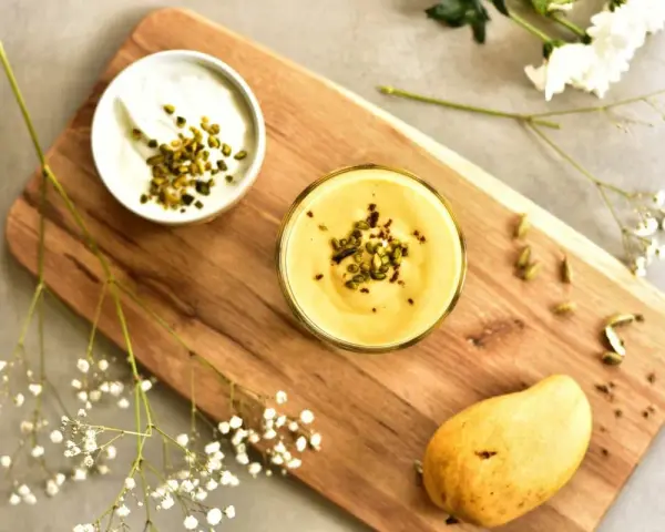 Aksamitny mango lassi w szklance, posypany pistacjami. Obok miseczka jogurtu, dojrzałe mango i kardamon.