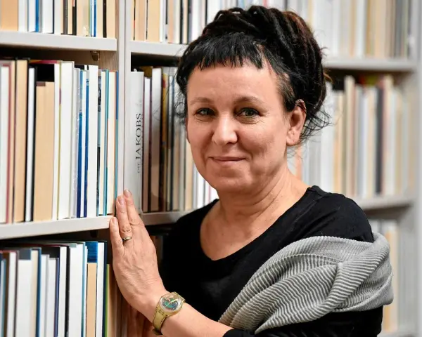 Olga Tokarczuk, w swoim wieku, otoczona książkami, z dłonią na grzbietach.
