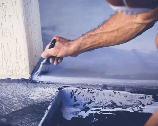 Mężczyzna maluje pędzlem szarą farbą betonową przy ścianie. Widać kuwetę z farbą.