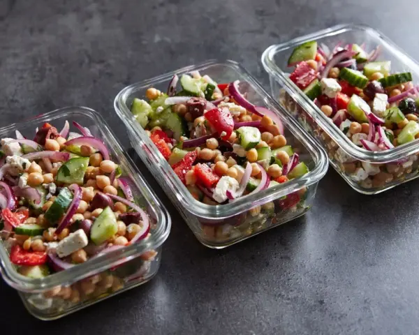 Trzy szklane pojemniki wypełnione kolorową sałatką z ciecierzycą, ogórkiem, pomidorem, czerwoną cebulą i fetą. Idealna na lunch.