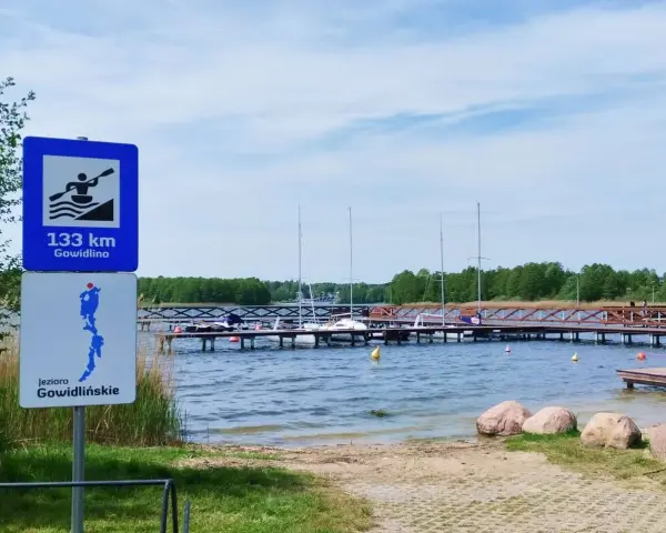 Znaki informują o 133 km do Gowidlina i o jeziorze Gowidlińskim. Widok na pomost z żaglówkami i kajakiem.