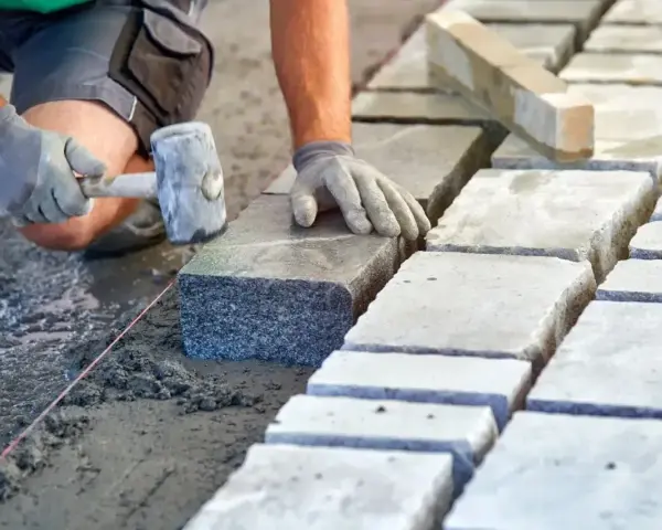 Ręce w rękawiczkach układają kamienne kostki na podsypce. Widać młotek i sznur geodezyjny. Prawidłowe suchy beton proporcje to klucz do trwałej nawierzchni.