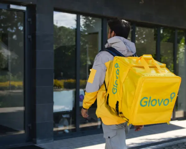Kurier Glovo pieszo idzie z żółtą torbą na plecach.