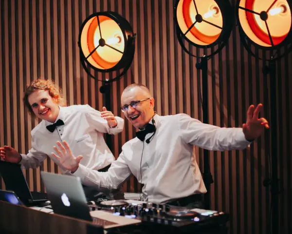 Dwóch DJ-ów w białych koszulach i czarnych muszkach, z uśmiechem na twarzy, przygotowuje się do imprezy. Zastanawiasz się, ile kosztuje DJ na wesele?