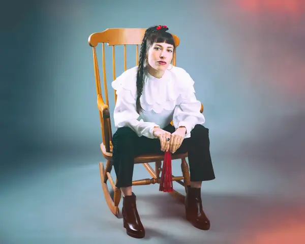 Scout Niblett, assise sur un fauteuil à bascule, porte une blouse blanche à volants et un pantalon noir. Elle tient un pompon rouge.