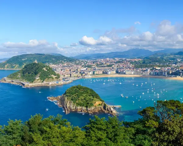 Widok na zatokę La Concha w Donostia San Sebastian, z zielonymi wzgórzami, miastem i licznymi łodziami na turkusowej wodzie.