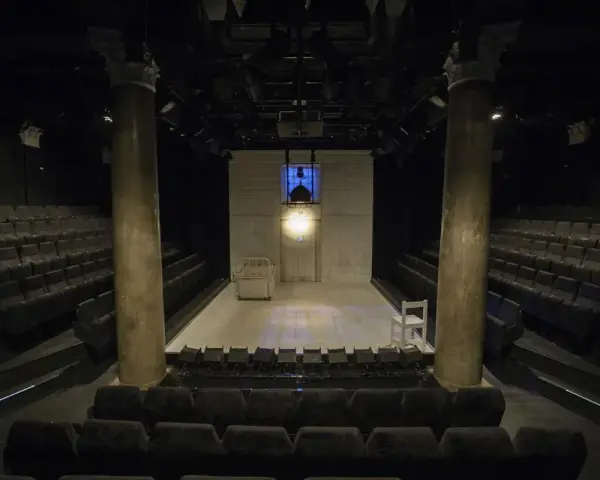 Pusta scena w **teatrze stu** miejsc, z rzędami siedzeń po bokach. Na scenie łóżko, białe krzesło i reflektory.