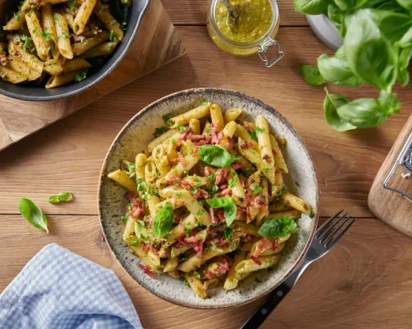 Makaron penne z pesto, boczkiem i bazylią na drewnianym stole.