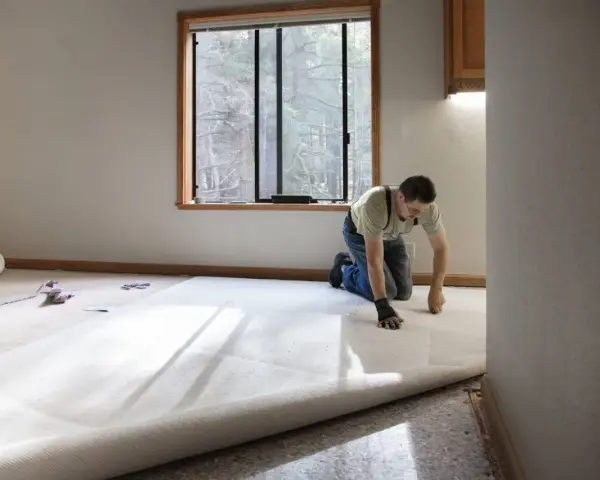 Un hombre instala una moqueta nueva en una habitación vacía. La moqueta es de color crema y se desenrolla por el suelo.