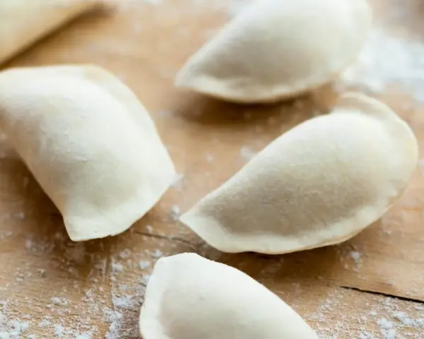 Surowe pierogi na drewnianej desce, przygotowane z ciasta na pierogi bez jajka.