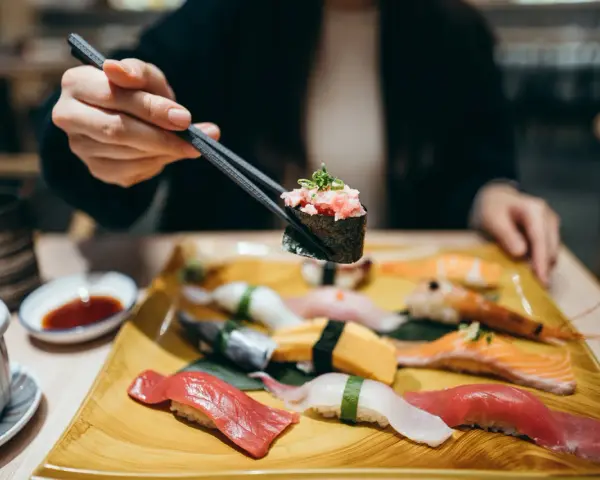 Eine Hand hält mit Stäbchen ein Stück Sushi mit gehacktem Thunfisch. Auf dem Teller liegen verschiedene Sushi-Rollen und Nigiri, bereit für den Genuss in Berlin.