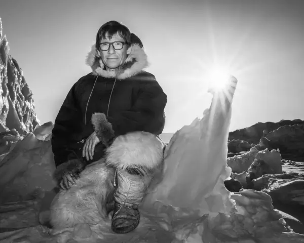 Ein Mann in traditioneller Inuit-Kleidung sitzt in eisiger Landschaft. Die Sonne scheint hell.