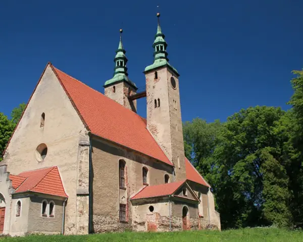 Stary kościół w stolcu z dwiema wieżami i zielonymi dachami, otoczony bujną zielenią drzew pod błękitnym niebem.