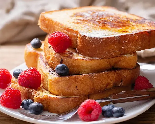 Una pila de tostadas francesas doradas, decoradas con frambuesas y arándanos, perfectas para aprender cómo se hacen las tostadas francesas.