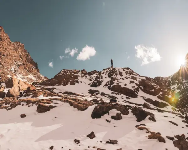 Ein Wanderer steht auf dem Gipfel des Jbel Toubkal, umgeben von schneebedeckten Felsen und einem strahlenden Sonnenschein.