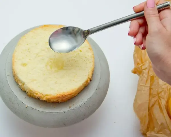 Dłoń z różowym lakierem do paznokci polewa łyżką biszkopt ponczem do tortu. Obok leży połówka cytryny.