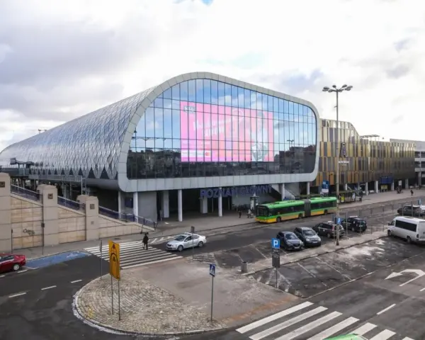 Nowoczesny Poznań Główny z charakterystyczną szklaną fasadą i ekranem. Autobusy i samochody czekają na pasażerów.