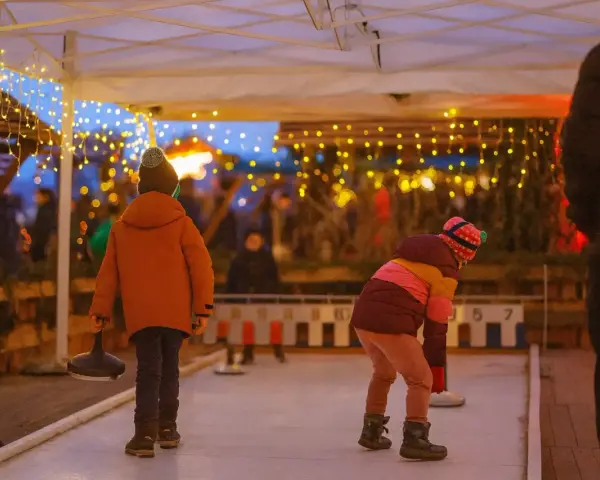Kinder spielen Eisstockschießen auf einem Weihnachtsmarkt in Berlin. Lichterketten leuchten im Abendlicht.
