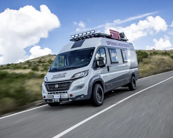 Szary kamper Ducato 4x4 Expedition z bagażnikiem dachowym i dodatkowym oświetleniem jedzie po drodze wśród wzgórz.