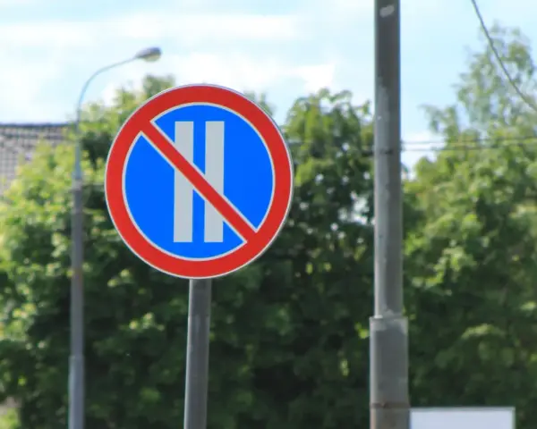 Znak B38 zakazujący postoju. Okrągły znak z czerwonym obramowaniem, niebieskim tłem i dwoma białymi pionowymi pasami przekreślonymi czerwoną linią.