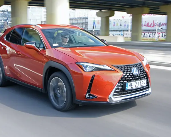 Pomarańczowy Lexus UX 250h jedzie po miejskiej drodze pod wiaduktem.