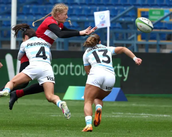 Zacięta walka o piłkę podczas rugby mistrzostwa świata. Zawodniczka w czerwonej koszulce próbuje przechwycić piłkę, podczas gdy dwie rywalki w białych strojach próbują ją zatrzymać.
