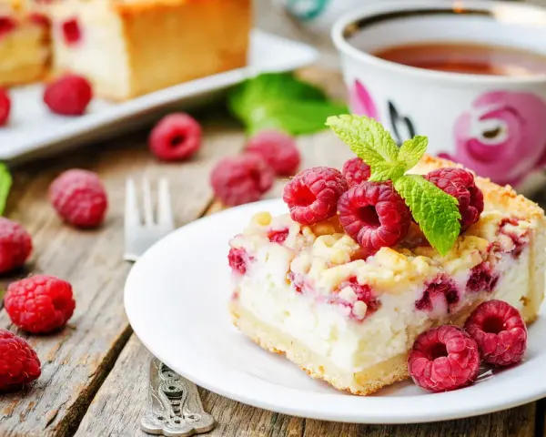 Malinowa chmurka z serem i malinami, udekorowana listkiem mięty. Obok filiżanka herbaty i świeże maliny.