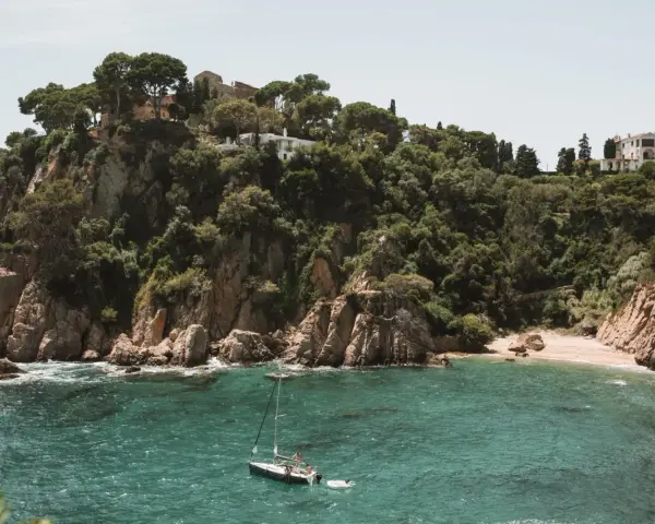 A sailboat floats in turquoise water near a rocky, tree-covered cliff with buildings. Perfect for weekend trips from Barcelona by train.