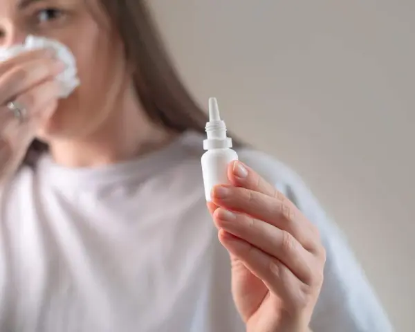 Kobieta z chusteczką przy nosie trzyma buteleczkę z kroplami do nosa, przygotowując się do plukania zatok.